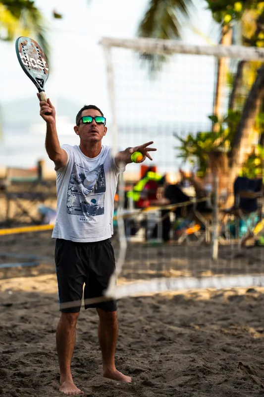 Un joueur de beach tennis lance la balle pour servir, pieds nus sur le sable.