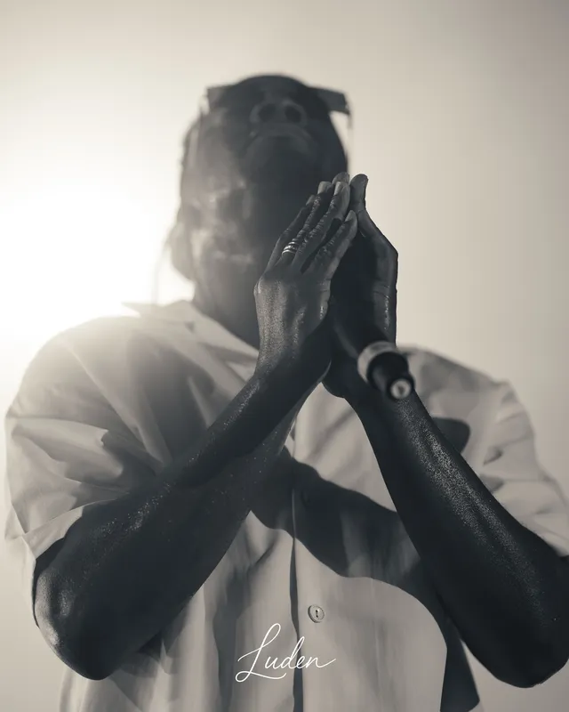 Un chanteur en contre-jour, les mains jointes devant le visage. Noir et blanc.