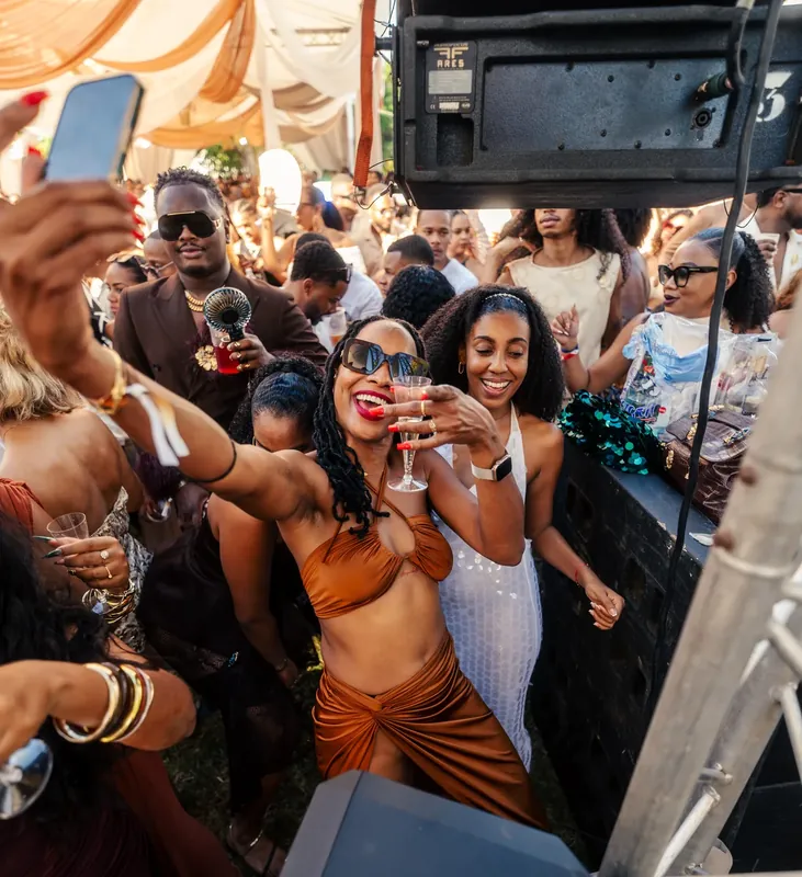 Des invités lèvent leurs verres et leurs téléphones sous un chapiteau aux draperies orangées.